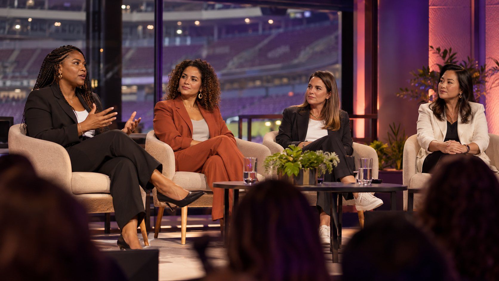 Quatro mulheres conversam sentadas em um palco, com plateia em primeiro plano e um estádio iluminado ao fundo.