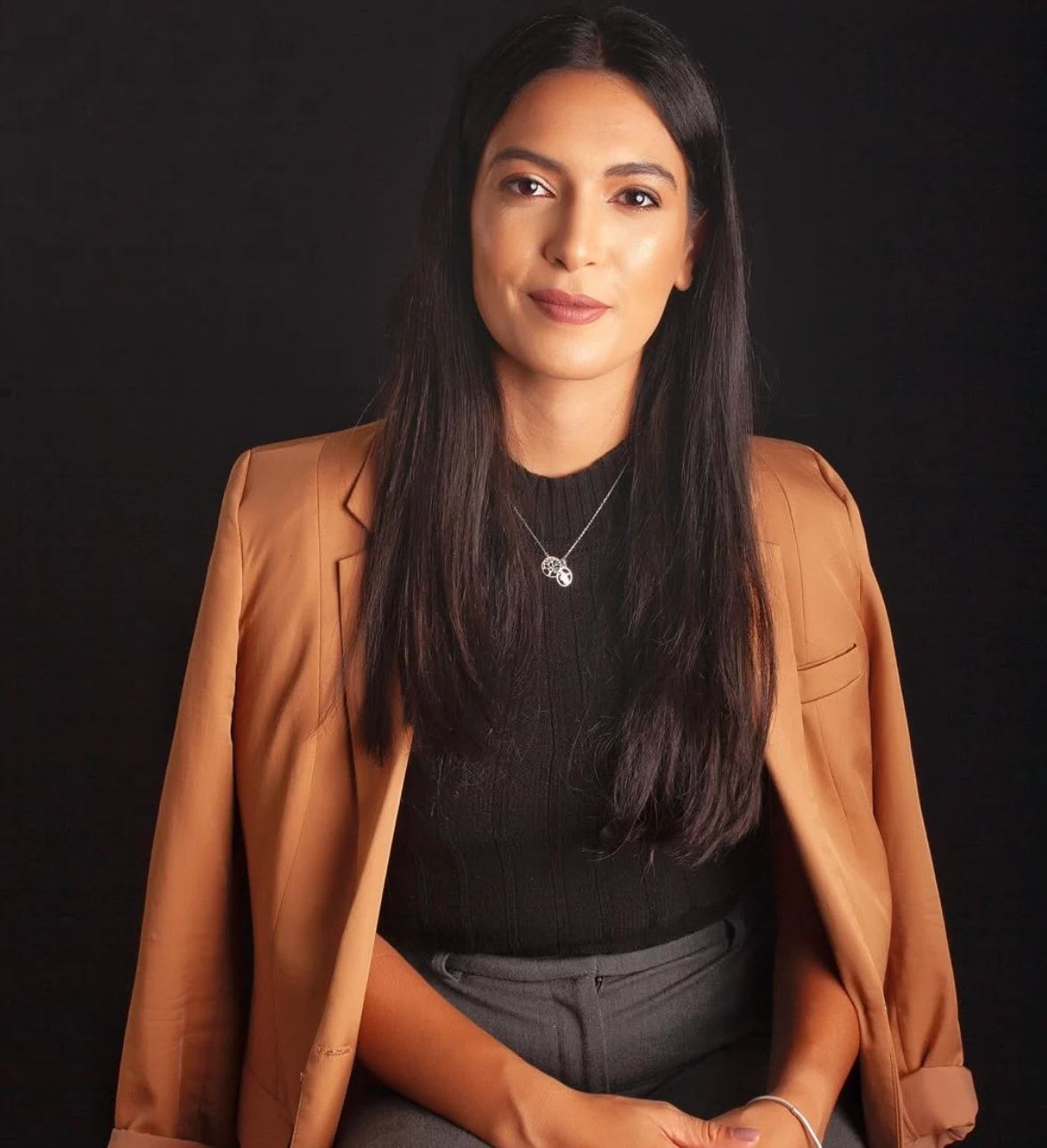 Retrato de Tamiris Monteiro, de cabelos longos e escuros, blazer caramelo sobre blusa preta, sentada diante de um fundo escuro.